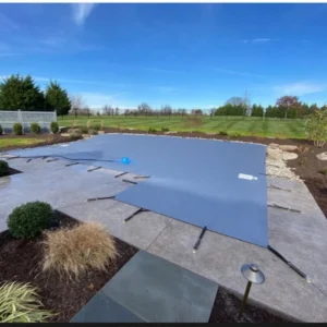A closed outdoor swimming pool is covered with a gray safety cover, surrounded by a concrete patio, landscaping, and a grassy field under a blue sky.
